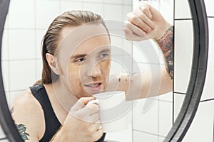 Unshaven man with patches under his eyes and cup of coffee looks at himself in bathroom mirror