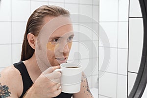 Unshaven man with patches under his eyes and cup of coffee looks at himself in bathroom mirror