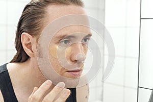 Unshaven man with patches under his eyes looks at himself in bathroom mirror