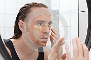 Unshaven man with patches under his eyes looks at himself in bathroom mirror