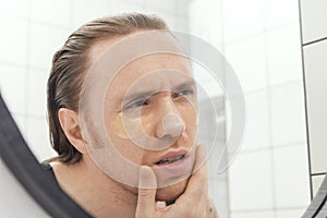 Unshaven man with patches under his eyes looks at himself in the bathroom mirror. Male with morning hangover