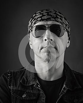 Unshaven, middle-aged man in a T-shirt, denim vest, bandana and sunglasses. Black and white portrait