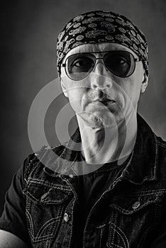 Unshaven, middle-aged man in a T-shirt, denim vest, bandana and sunglasses. Black and white portrait