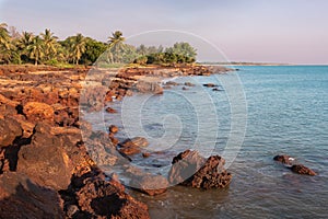 Unspoiled beach at sunset time. Rocky landscape, green trees and palm trees. Coastline of Timor sea. No people in the picture.