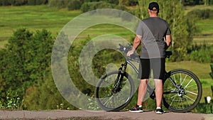 Unsportsmanlike overweight man with his mountain bicycle rests after riding. Cyclist is standing on hill against