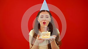 Unsuccessful attempt to blow out the candle and make a birthday wish. Young woman in a festive cap holds a plate with a slice