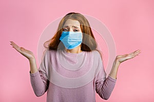 Unsure woman in medical mask shrugs her arms, makes gesture of I don`t know, care, can`t help anything . Girl on pink
