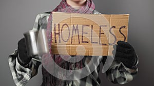 A untidy beggar woman in dirty clothes and gloves begging and holds a cardboard sign with the inscription homeless. Dark