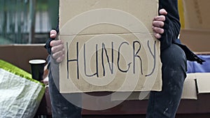 An untidy man sits on the street and holds a handwritten HUNGRY poster.