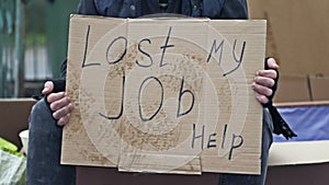 An untidy man is sitting with a cup of some drink and holding a handwritten LOST MY JOB HELP placard.