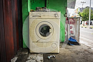 The unused white washing machine with front door near green wall and a broom photo abandoned in a street photo taken in