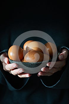 Unusual Easter on dark background. Bowl of brown eggs with hands, vertical