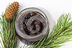 Unusual jam from pine cones in glass jar on white background among pine branches and cones. Organic and vegetarian sweet dessert