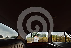 Unusual view of stylish bride and happy groom through red retro car on the background of nature