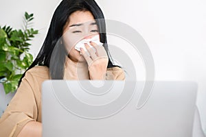 Unwell Asian woman sneezing and coughing because of flu and fever while working on computer at desk hand holding tissue papers