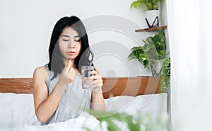 Unwell Asian woman taking medicine with glass of water sitting in bed