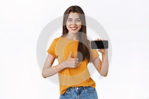 Upbeat, satisfied good-looking assertive woman in yellow t-shirt, showing smartphone display horizontally and thumbs-up