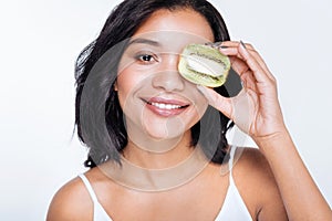Upbeat woman holding kiwifruit near her eye