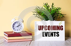 UPCOMING EVENTS is written in a notebook next to a green plant and a white alarm clock, which stands on colorful diaries