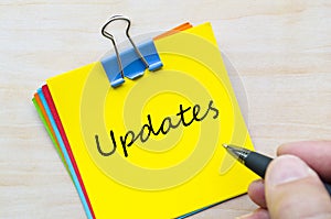 Updates handwritten text on yellow notepad clipped with silver pin on light wooden desk.