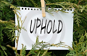 UPHOLD - word on white paper with clothespin on a green background of branches