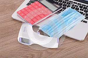 Upper mask, infrared thermometer and calculator on laptop keyboard