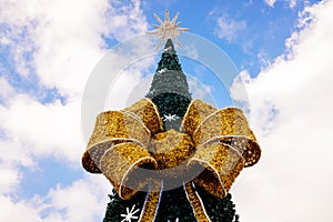Upper part of a large Christmas tree adorned with a large golden bow, in the open sky with clouds