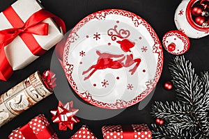 Upper top view of a red ribbon, Christmas presents, tree toys, deer plate and evergreen branch on a stone black background.