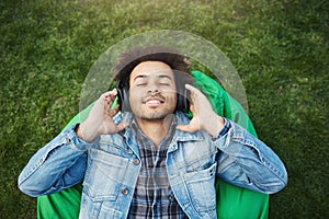Upper view portrait of pleased relaxed african-american man with bristle lying on grass while listening music with