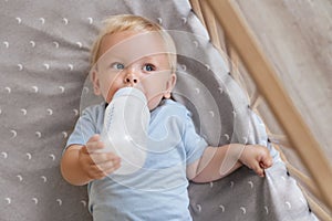 Upper view portrait of sweet baby lying in bed feeding himself holding bottle with formula