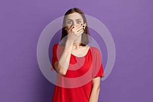 Ups, I said too much. Confused young pretty brunette girl in red t-shirt covering her mouth with palm like she told something bad