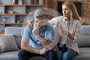 Upset adult caucasian wife calm husband, gives glass of water, man suffers from pain and depression