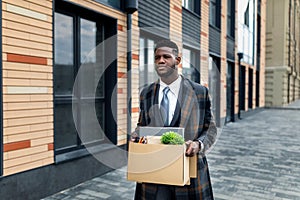 Upset african american male office worker holding box with things and leaving office building, copy space