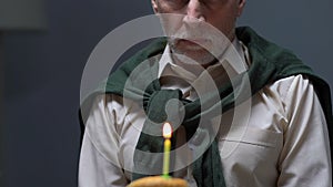 Upset aged man sitting alone with birthday cake on table, abandoned by family