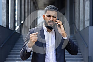 Upset and angry Indian male businessman standing outside an office center and talking on the phone emotionally