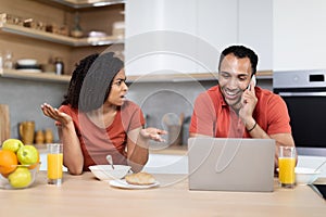 Upset angry millennial black female swears at man, speaks on phone with laptop in kitchen interior