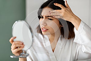 Upset arabic woman looking at hand mirror, checking skin