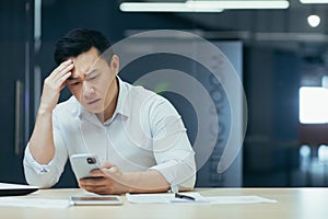 Upset asian businessman working in office, reading bad news on phone, man depressed at work