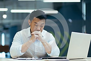 Upset Asian male businessman sitting in the office at the table, thoughtfully leaning his head on his hands. Financial