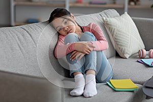 Upset asian schooler sitting on couch, studying at home