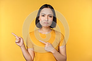 Upset asian woman pointing fingers left, showing smth disappointing, complaining at banner, standing over yellow