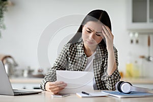 Upset asian woman sitting in kitchen, reading letter