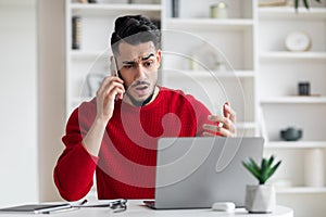 Upset attractive millennial arabic middle eastern in red clothes call by phone, gesticulate, look at laptop in home