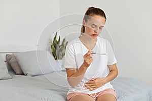 Upset attractive woman wearing casual attire sitting on sofa at home and holding pregnancy test with positive result, holding her