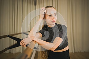 Upset ballerina in training suit stands near ballet barre and looks at the mirror in dance studio. Woman prepares for