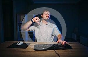 An upset bearded man works at night on a computer at home and shows a dislike gesture. Mirthless freelance programmer shows thumbs