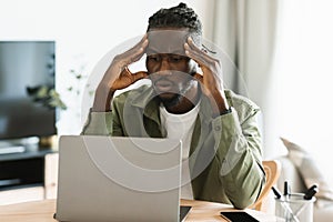 Upset black freelancer man sitting at workdesk at home office, working on laptop and suffering from headache