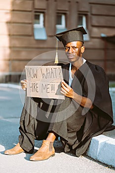Upset black guy sitting with cardboard poster on street looking for job. University or college graduating student in