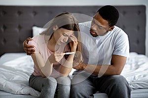 Upset black man comforting his depressed Caucasian wife with negative pregnancy test in bedroom