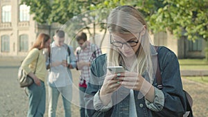 Upset blond girl scrolling smartphone screen as blurred groupmates laughing at the background. Nerd young Caucasian